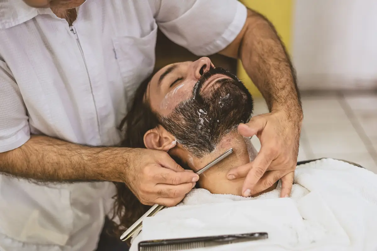 A barber shaves a reclining, bearded man's jawline with a straight razor, a white towel across his chest