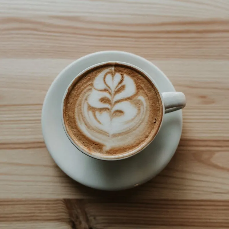 A latte with leaf art in a plain white cup and saucer on a wooden table