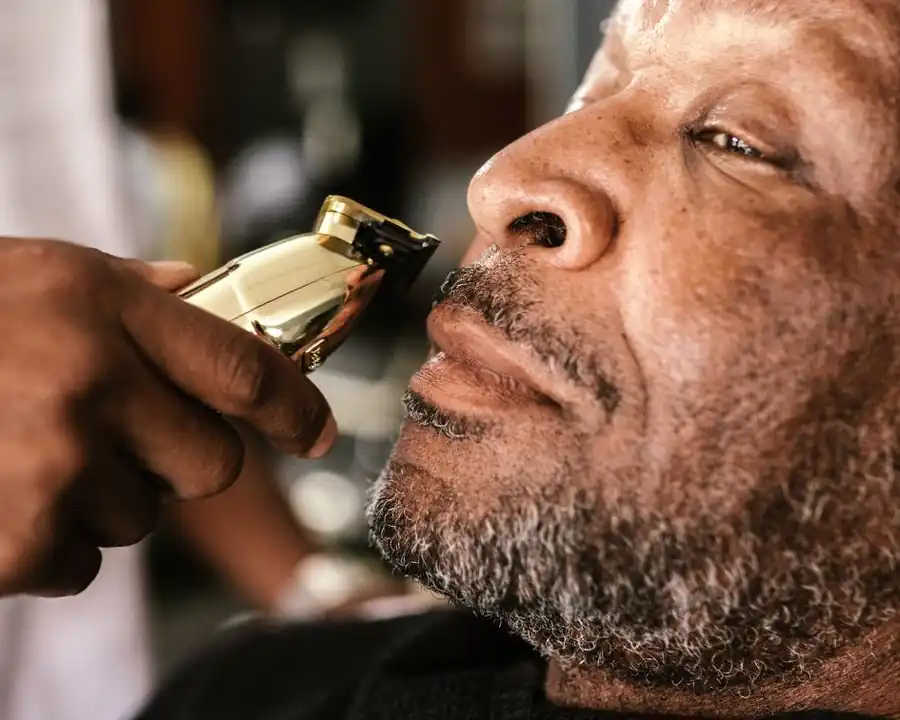 A barber lines up an older man's grey moustache with a gold trimmer