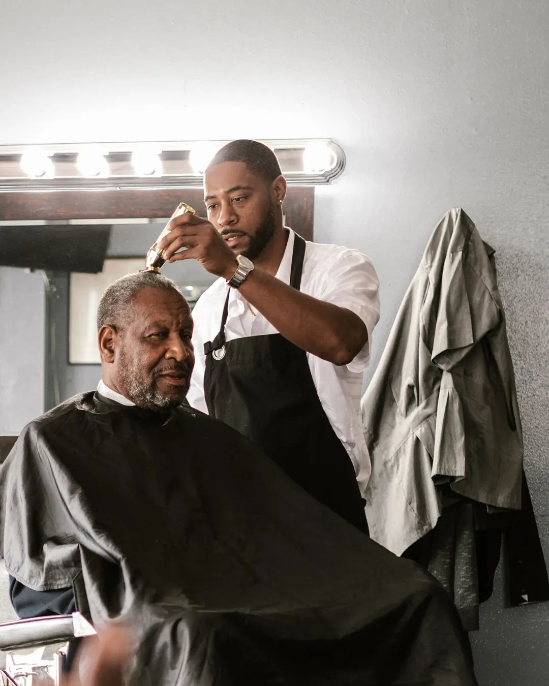 A barber in a black apron clips an older client's grey hair in front of a bulb-lit mirror